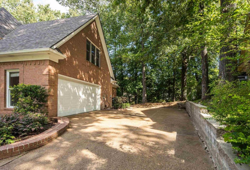 Brick exterior featuring a two-car garage with a white door, a sloped concrete driveway, a brick retaining wall, and mature trees