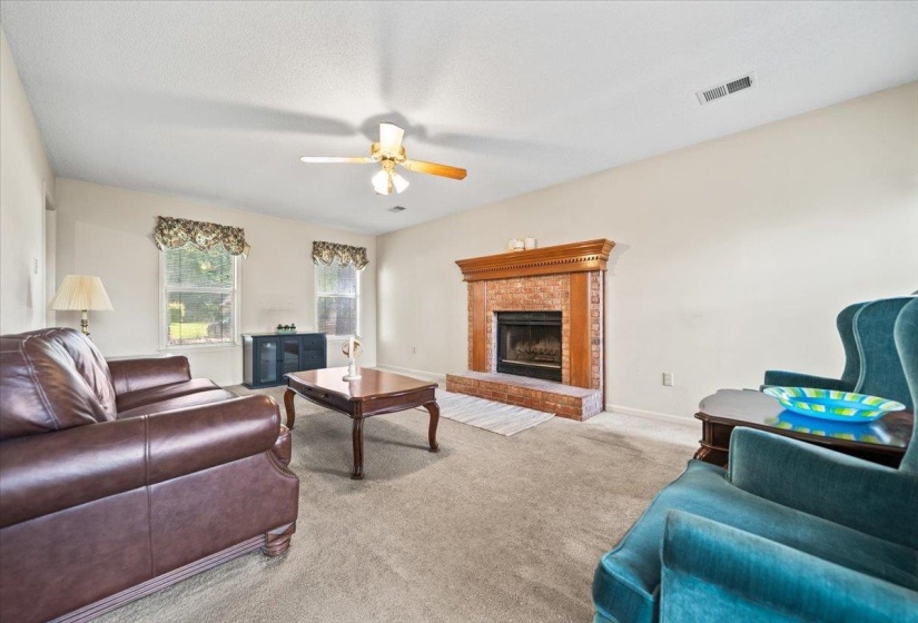 Living area featuring a brick fireplace with a wood-finish mantel, neutral carpet flooring, and a ceiling fan