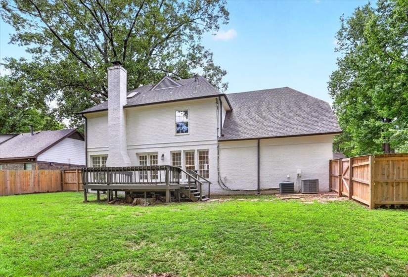Spacious backyard featuring a large elevated deck with railing, a prominent brick chimney, multiple ground-level windows, and a privacy fence
