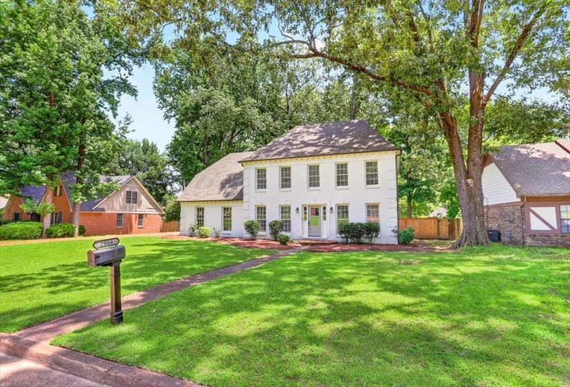 Traditional two-story residence with a white brick exterior, dark roof, and mature tree landscaping