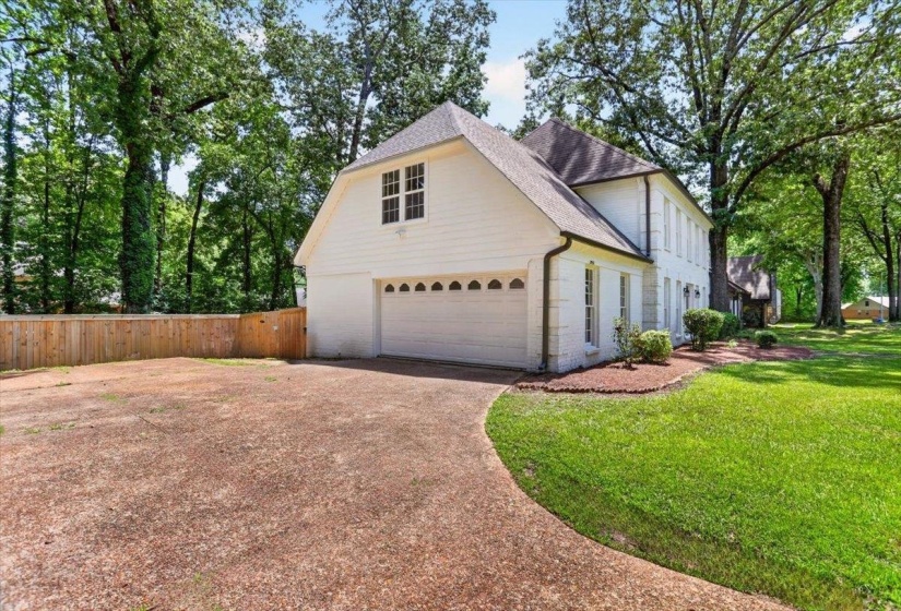 White exterior featuring a two-car garage with decorative window inserts, an expansive gravel driveway, a wooden privacy fence, mature trees, and a manicured lawn