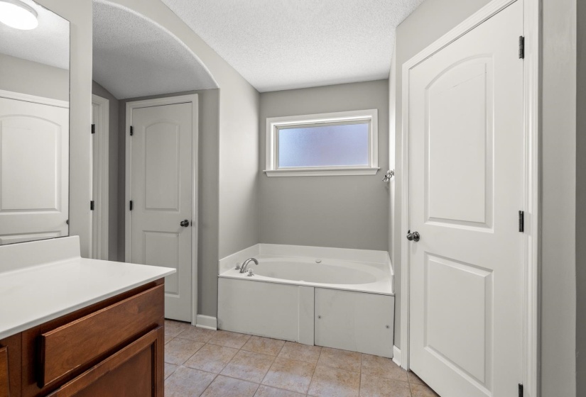 Soaking tub with a frosted privacy window, complemented by a wood-finish vanity with a white countertop, tile flooring, and arched doorway