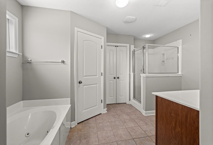 Spacious bathroom featuring a built-in jetted tub, a glass-enclosed shower, a wood-finish vanity with a light-toned countertop, and durable tile flooring