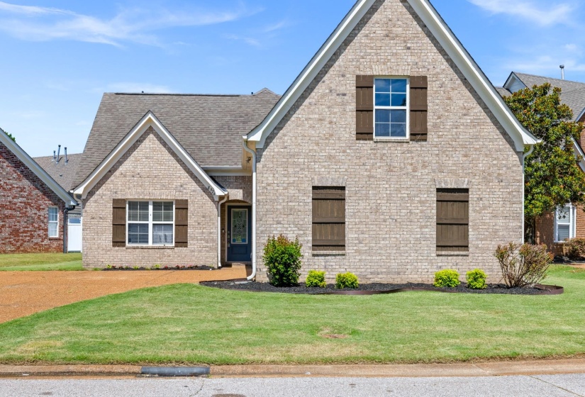 Brick exterior residence featuring a covered entry, dark wood shutters, gable rooflines, an attached garage, and landscaping with shrubs and manicured lawn