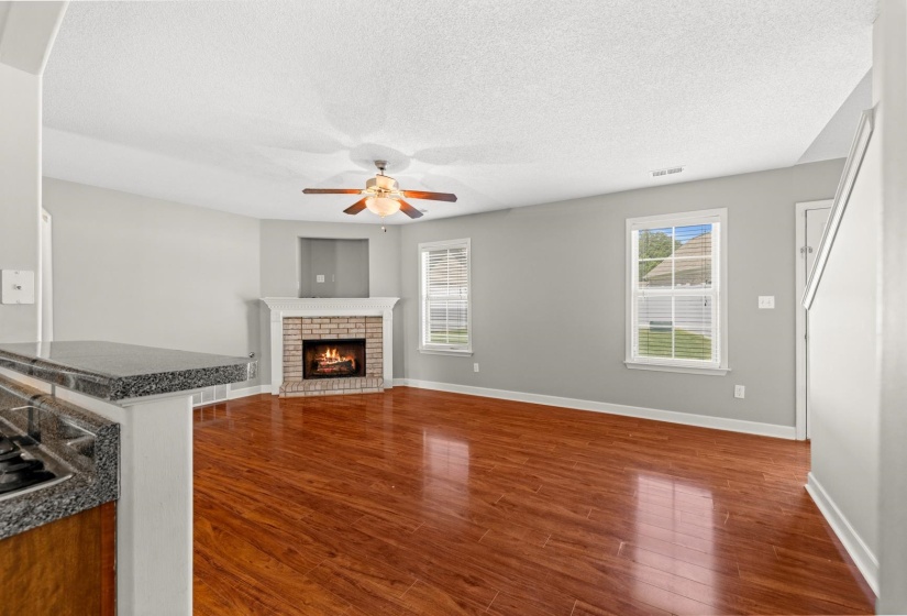 Spacious living area featuring wood-finish flooring and a corner fireplace with a brick surround and white mantle