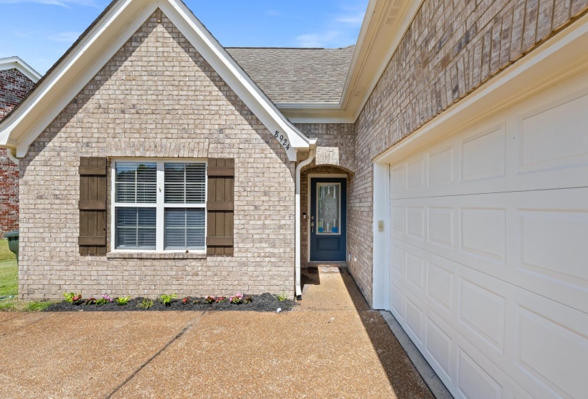 Brick exterior with a prominent gable, white-paned window with louvered shutters, decorative dark entry door, and an attached garage with a panel-style door