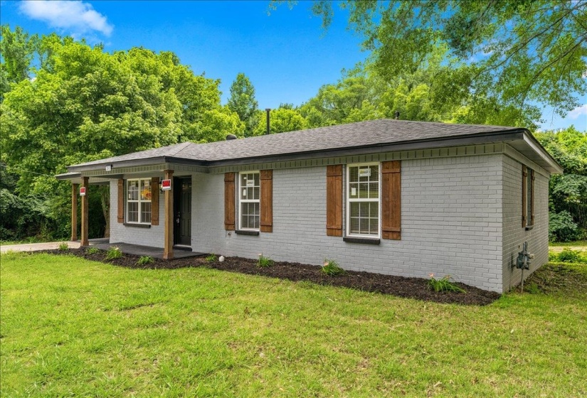 Gray brick exterior with dark wood shutters, a covered porch with wood columns, a dark shingle roof, and a landscaped front planter bed