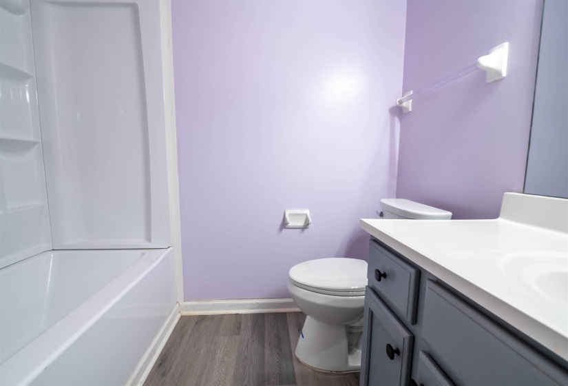 Bathroom featuring a white tub/shower combination, light purple walls, a toilet, a vanity with a white countertop and gray cabinetry, and wood-finish flooring
