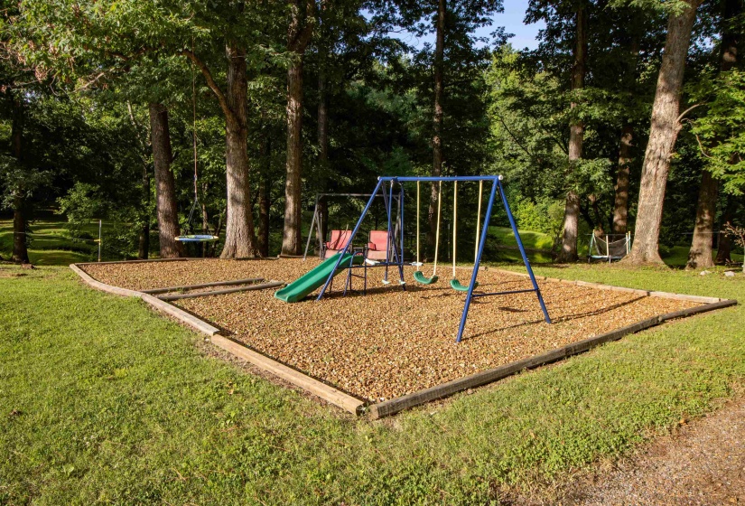 Outdoor play area featuring a swing set with slide, a separate tire swing, and a wood chip surface, all surrounded by mature trees and a grassy lawn