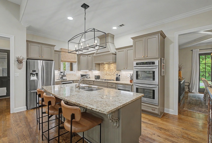 Gourmet kitchen featuring a large central island with integrated sink, stainless steel appliances, custom cabinetry, tiled backsplash, and wood-finish flooring