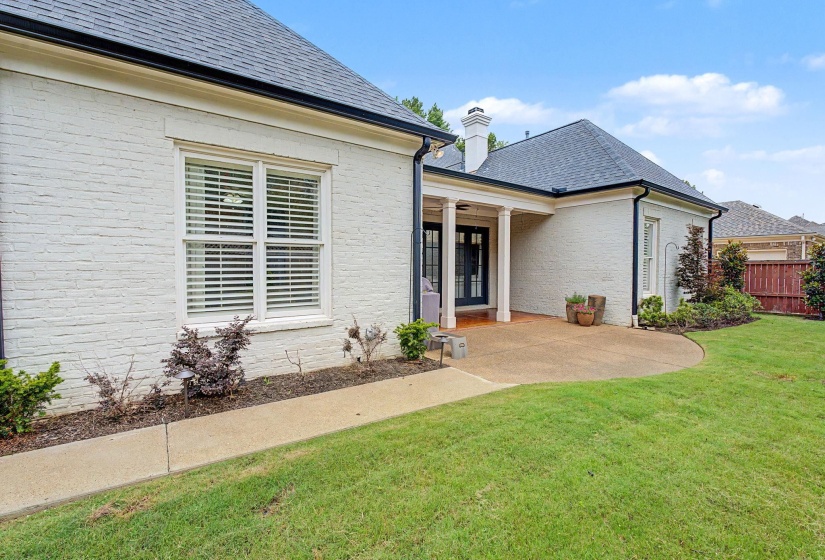Exterior featuring painted brick siding, dark-toned roof, covered patio with white columns, concrete patio, and a manicured lawn