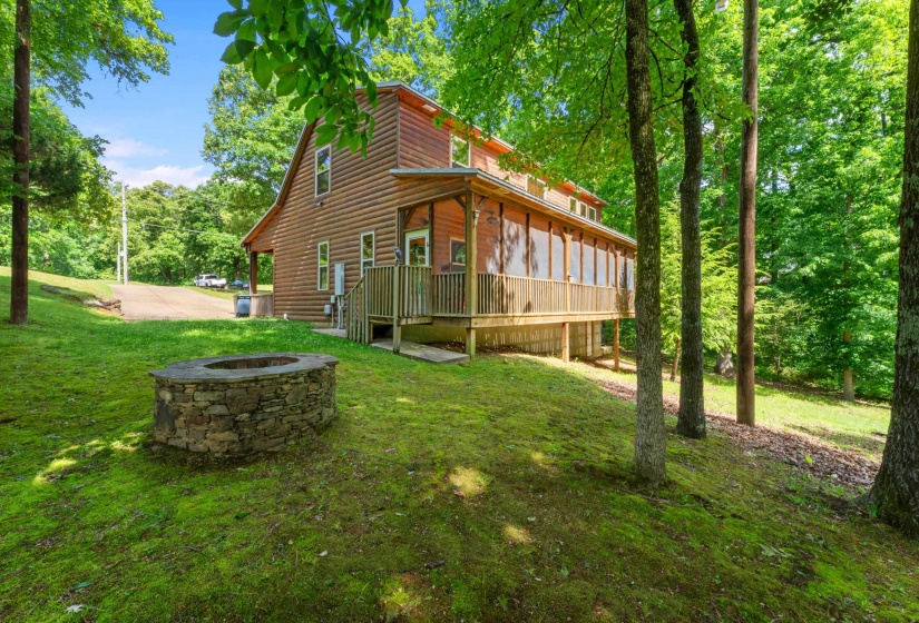 Two-story log cabin exterior featuring a covered front porch with railings, a screened-in back porch with wood railings, a stone fire pit, and a sloped, tree-lined lawn