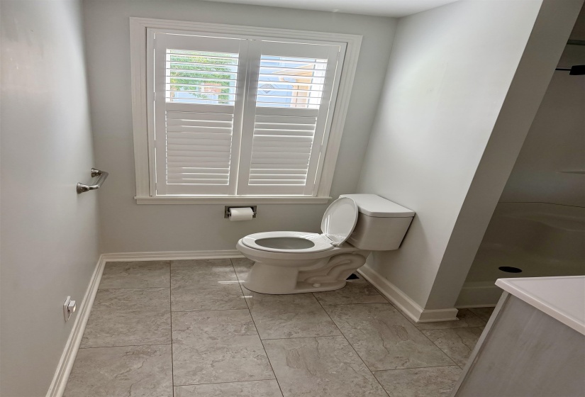 Bathroom featuring a window with white louvered shutters, light gray walls, and gray tile flooring
