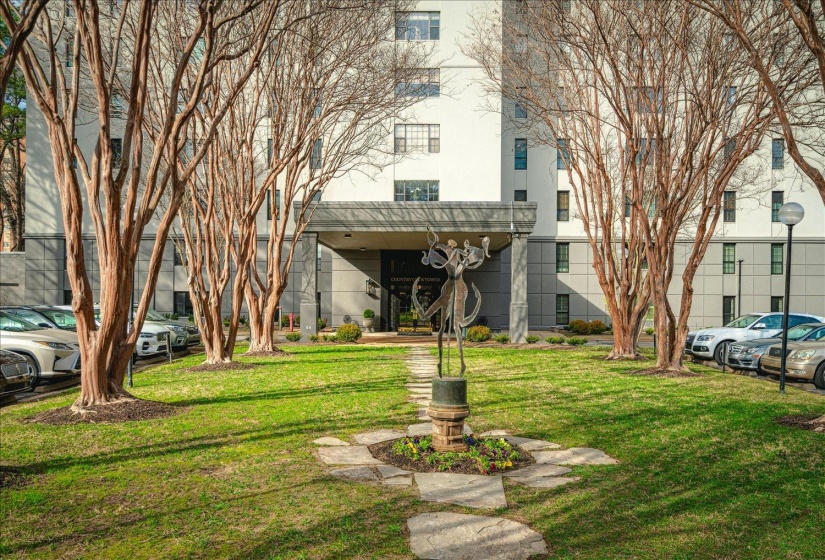 Modern high-rise building with a grand entrance canopy, flanked by mature deciduous trees and a landscaped lawn