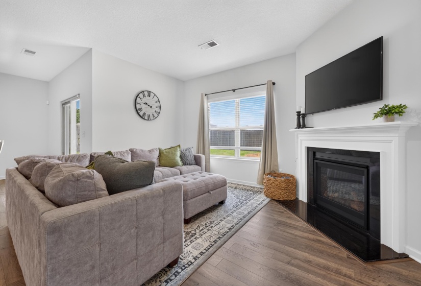 Living area featuring wood-finish flooring, a white mantel fireplace, and a large window with blinds
