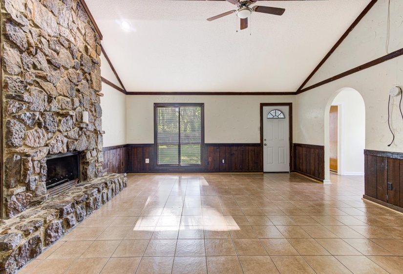 Spacious room featuring a stone-clad fireplace, vaulted ceiling with wood beams, tiled flooring, wood paneling, and an arched doorway