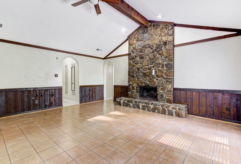 Vaulted ceiling with exposed wood beams, stone fireplace with hearth, tile flooring, and wood paneling