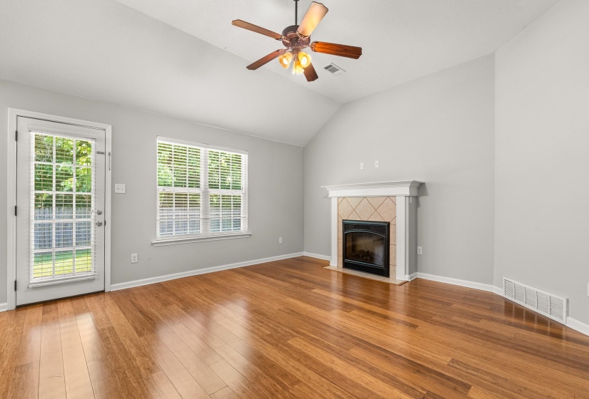 Spacious living area featuring wood-finish flooring, a fireplace with a decorative tile surround and white mantel, and a vaulted ceiling with a ceiling fan