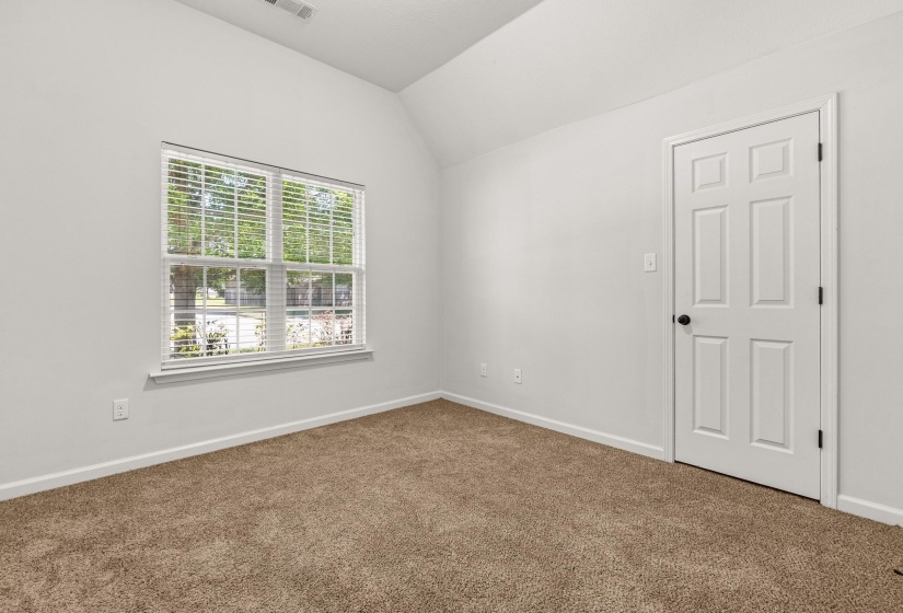 Neutral-toned bedroom featuring a vaulted ceiling, light beige carpeting, a large window with white blinds, and a paneled interior door with matte black hardware