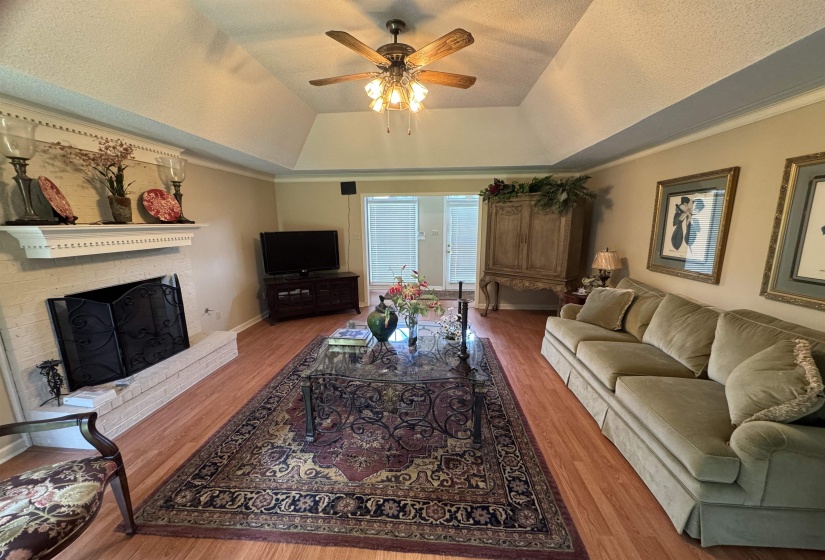 Living area featuring wood-finish flooring, a white brick fireplace with a decorative mantel, and a tray ceiling with a ceiling fan