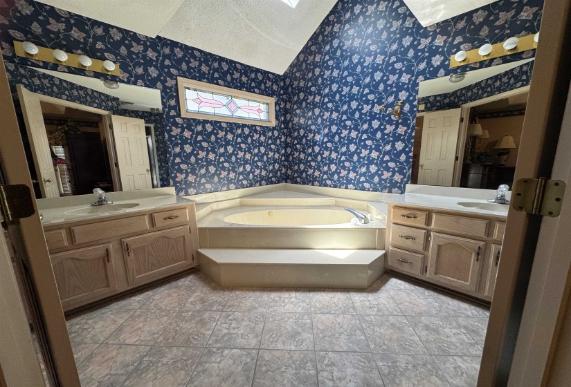Corner jetted tub with integrated steps, flanked by two vanities featuring light wood-finish cabinetry, white countertops, and integrated sinks
