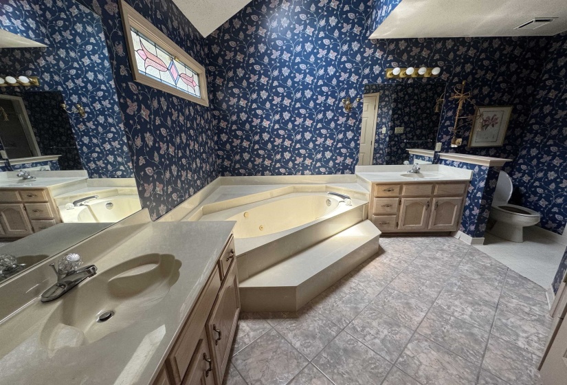Bathroom featuring a built-in jetted tub with integrated steps, dual vanity areas with undermount sinks, patterned wallpaper, and a stained glass window detail