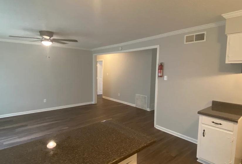 Open-concept living area featuring wood-finish flooring, a ceiling fan with integrated lighting, crown molding, and a pass-through counter with a dark countertop