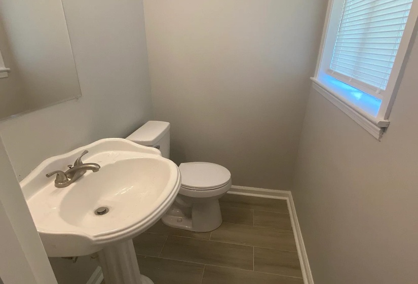 Pedestal sink with brushed nickel faucet, complete with a white toilet, wood-finish tile flooring, and a window with blinds