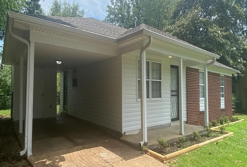 Exterior showcasing a covered carport, white vinyl siding, brick facade, a front porch, and mature trees in the background