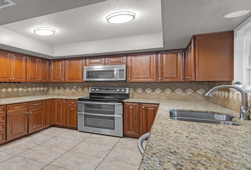 Kitchen featuring wood cabinetry, stainless steel appliances, granite-finish countertops, tile flooring, and a patterned tile backsplash