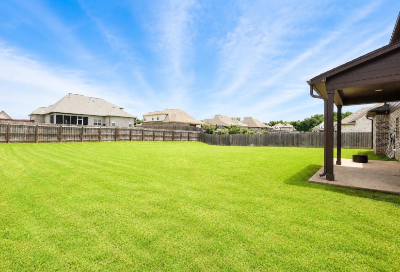 Expansive backyard featuring a large green lawn, full privacy fencing, and a covered patio with concrete flooring