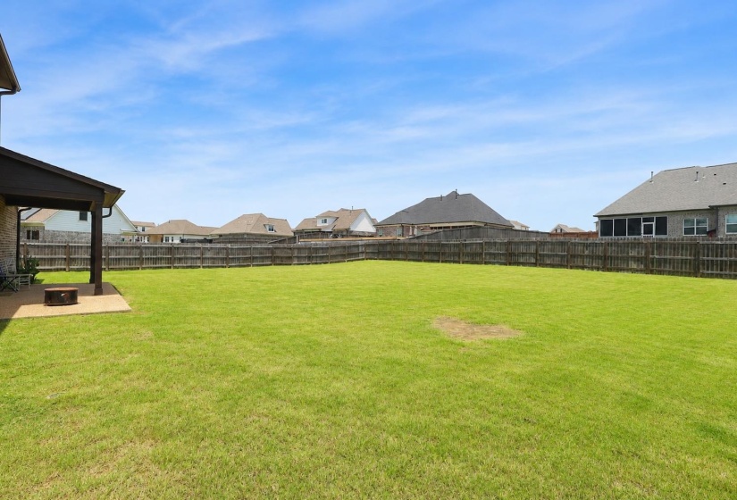 Expansive backyard featuring a large green lawn, full privacy fencing, and a covered patio area with concrete flooring and a built-in fire pit