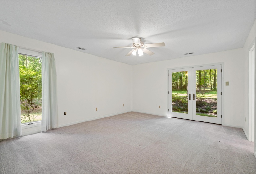 Spacious room featuring light carpeting, white walls, a ceiling fan, a large picture window, and double French doors opening to outdoor greenery