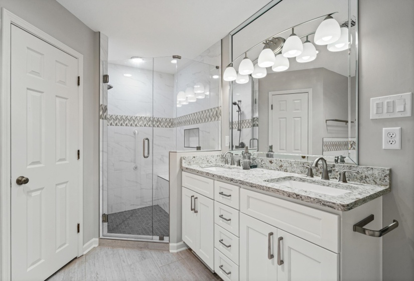 Bathroom featuring a double vanity with stone countertops, a large mirror with integrated lighting, a glass-enclosed shower with a built-in bench, a decorative tile accent band, and wood-finish flooring