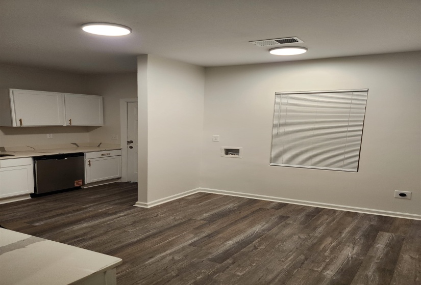 Open-concept living space featuring wood-finish flooring, white cabinetry, a stainless steel dishwasher, and a window with blinds