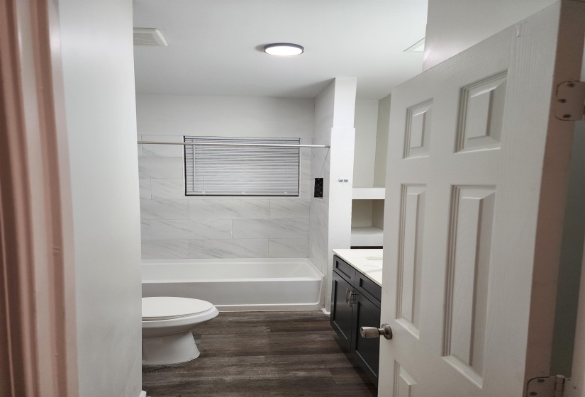 Full bathroom featuring a white tub with subway tile surround, wood-finish flooring, a dark-toned vanity with a light countertop, and built-in shelving