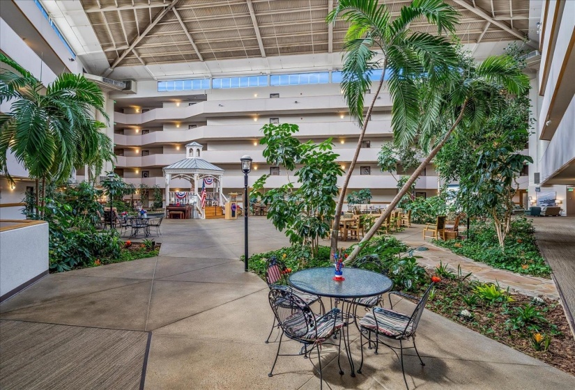 Multi-story atrium featuring a central gazebo structure, various seating areas, and extensive indoor landscaping with mature palm trees