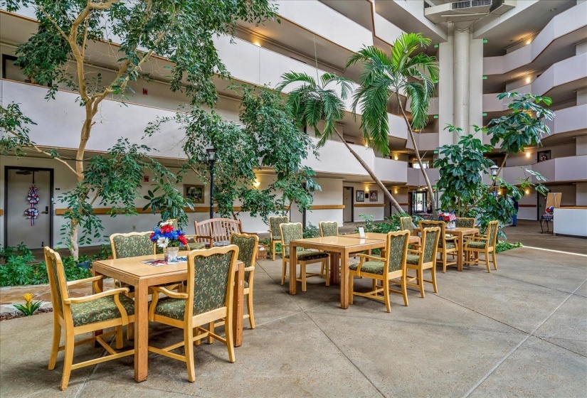 Spacious multi-story atrium featuring a combination of mature trees and tropical palm trees, multiple seating areas with wooden tables and upholstered chairs, and multi-level open-air corridors