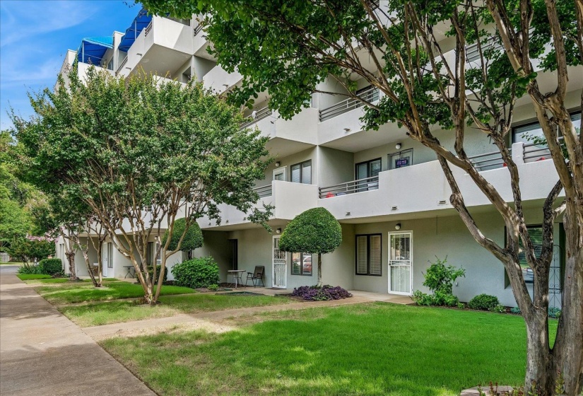 Multi-story building featuring private balconies, individual entryways, mature trees, and a concrete walkway