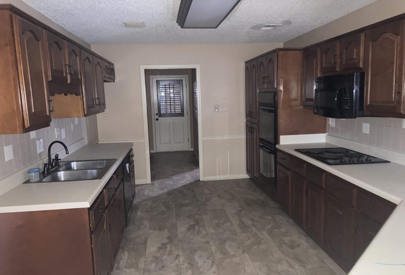 Galley kitchen featuring dark wood cabinetry, light countertops, a double basin stainless steel sink, a built-in oven, and a black cooktop