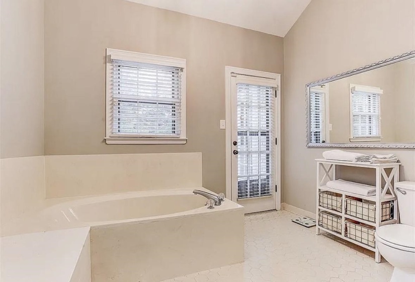 Soaking tub with deck surround, large window with blinds, full-lite door with blinds, textured tile flooring, and vaulted ceiling