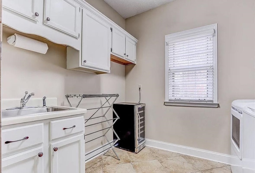Laundry room featuring white cabinetry, a built-in sink with a chrome faucet, a window with blinds, and tile flooring