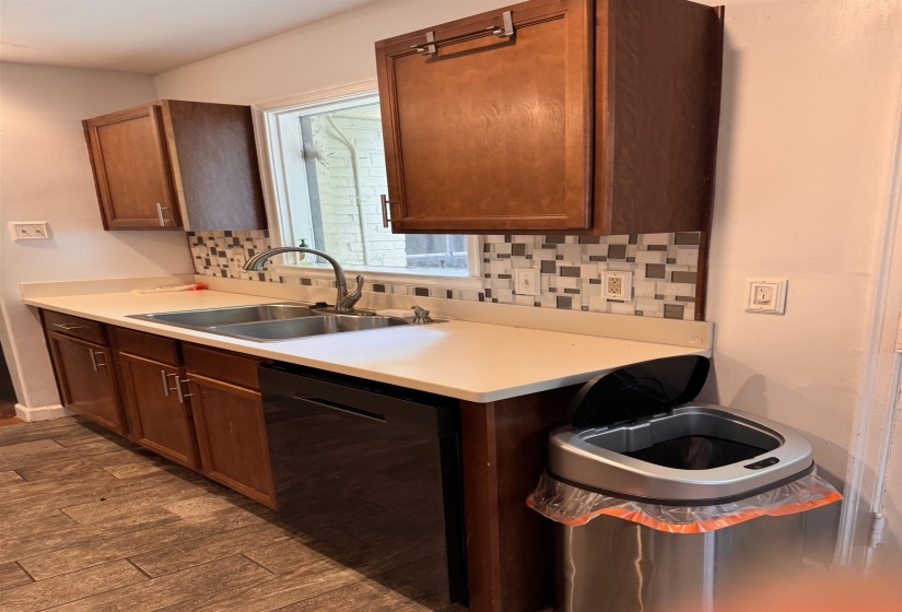Kitchen workstation featuring wood cabinetry, light-toned countertops, a stainless steel double-basin sink with a gooseneck faucet, a patterned tile backsplash, and a built-in black dishwasher