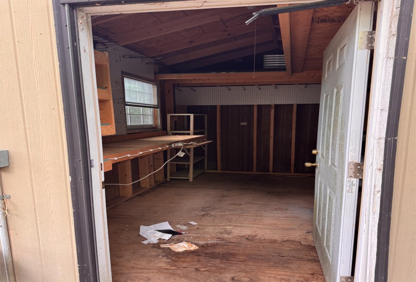 Utility shed interior featuring exposed wood-beam ceiling, plywood flooring, built-in workbench, and pegboard wall