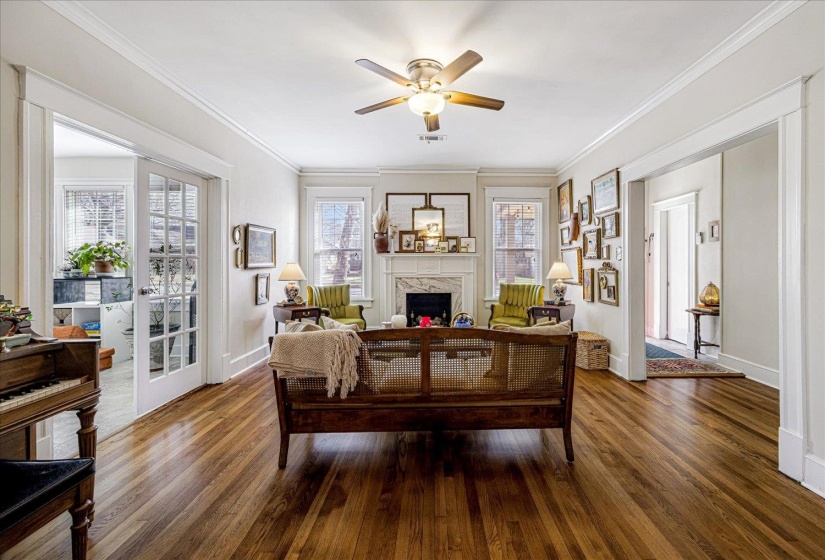 Living area featuring rich wood-finish flooring, a white mantel fireplace, multiple windows with natural light, a ceiling fan with light fixture, and French doors leading to an additional room