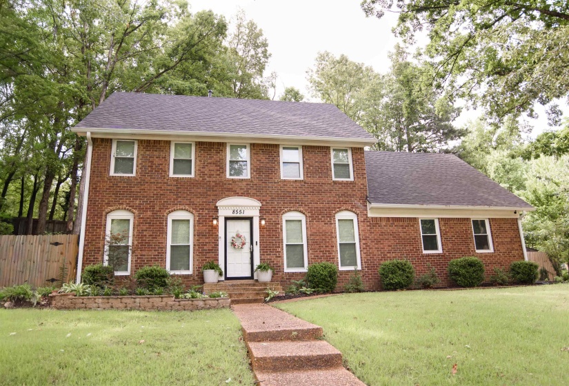 Traditional brick exterior featuring a white entry door, arched window details, multiple windows, dark shingle roof, and a stone-lined walkway