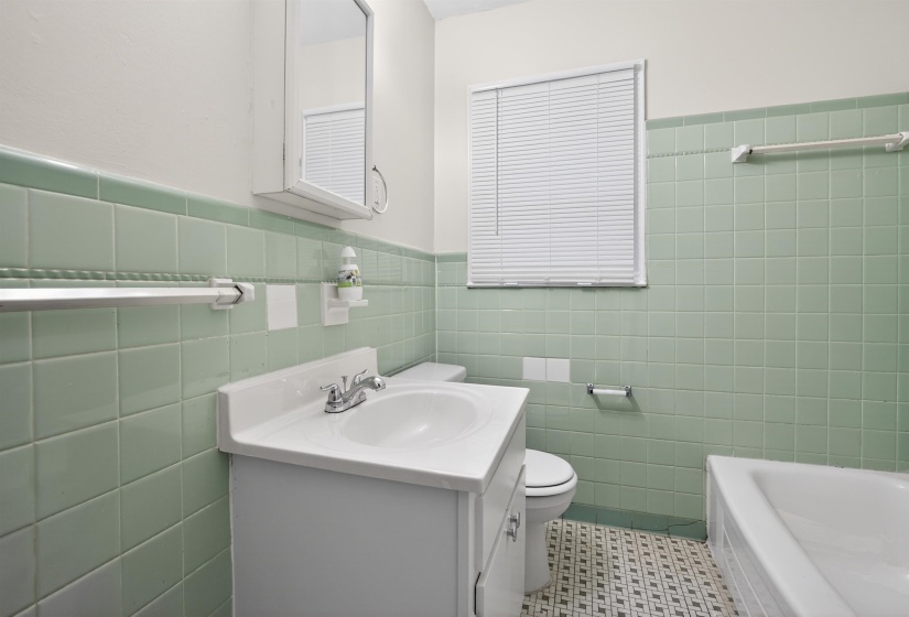 Bathroom featuring green wall tiling, a white vanity with an integrated sink, a built-in medicine cabinet, a window with blinds, and geometric floor tiling