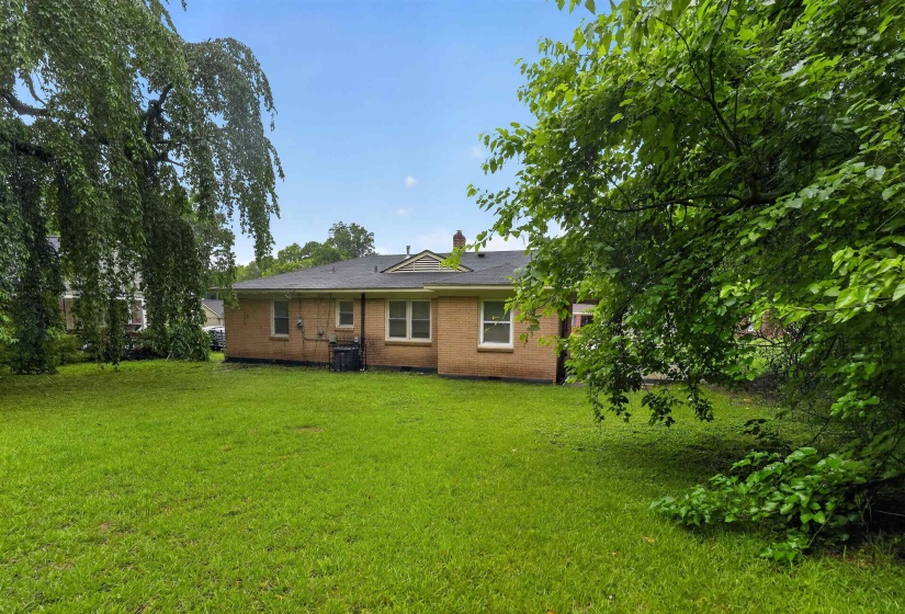 Expansive green lawn surrounds the rear of this property, featuring a brick exterior with white trim windows and a dark shingle roof