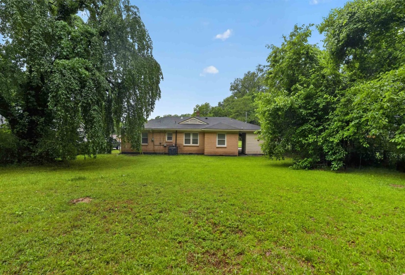 Expansive rear lawn featuring mature trees and a brick exterior home with multiple windows, black shingle roofing, and an attached white structure