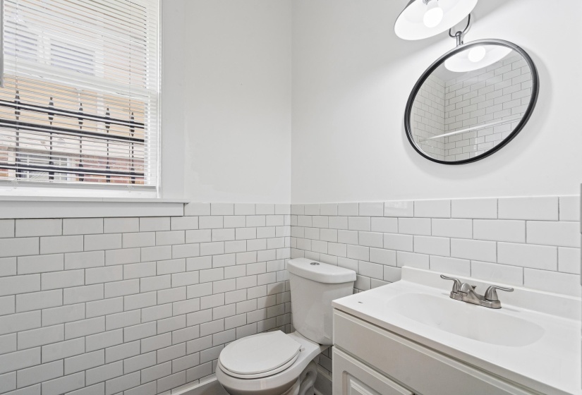 Bathroom featuring white subway tile wainscoting, a wall-mounted vanity with integrated sink, a circular mirror, and a window with blinds
